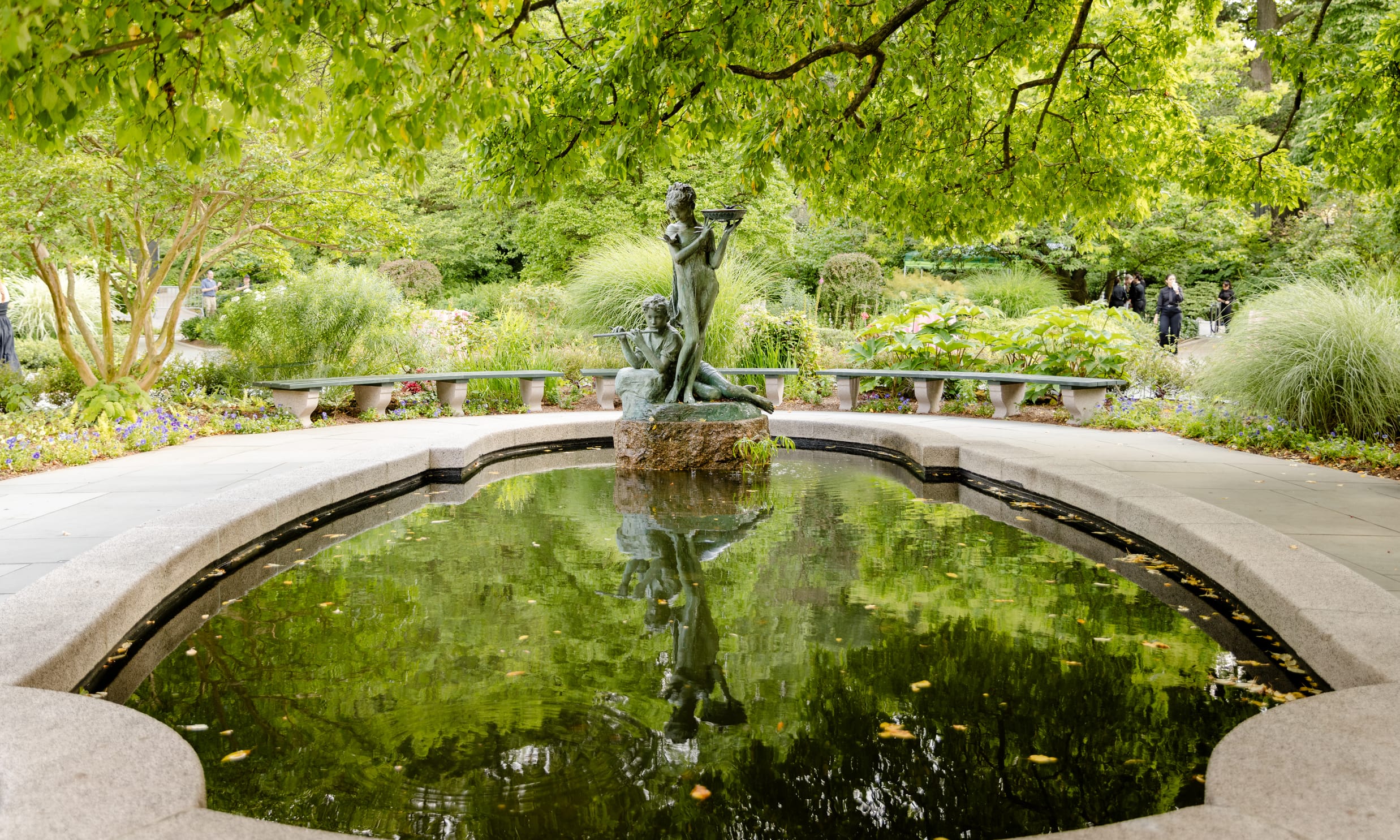 A green statue of two women stands over a small reflecting pool under a canopy of trees.