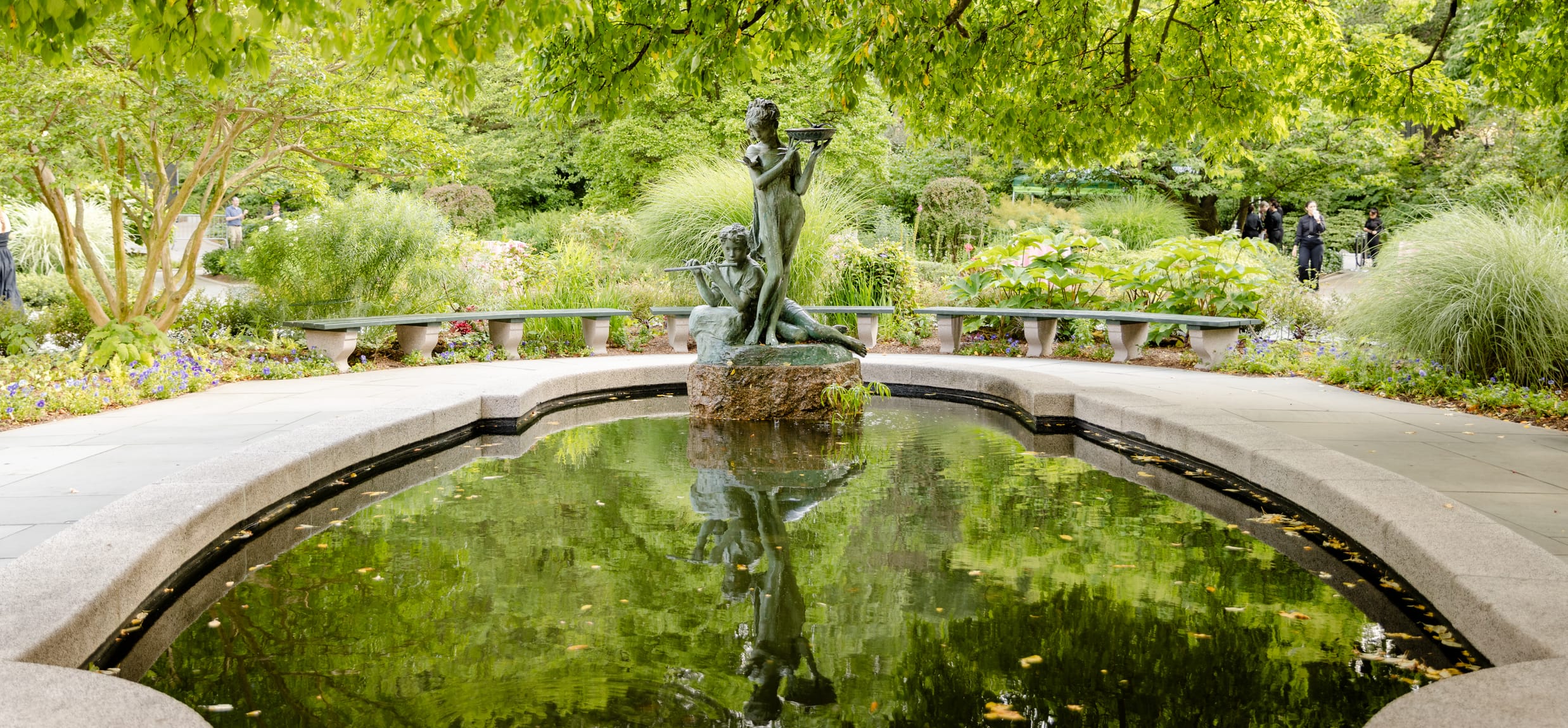 A green statue of two women stands over a small reflecting pool under a canopy of trees.