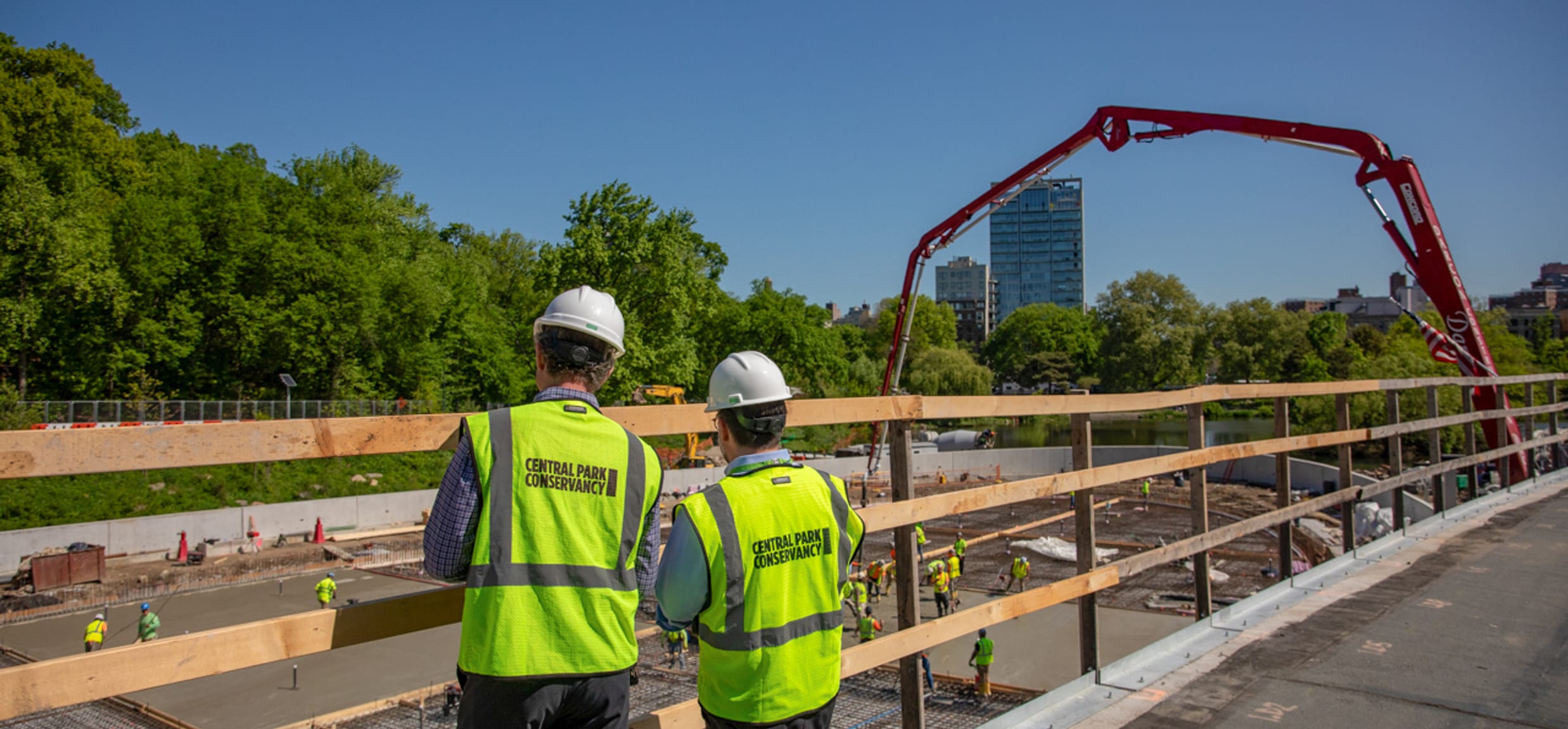 Two people stand together, overlooking a construction site, wearing hard hats and yellow vests reading “Central Park Conservancy.”