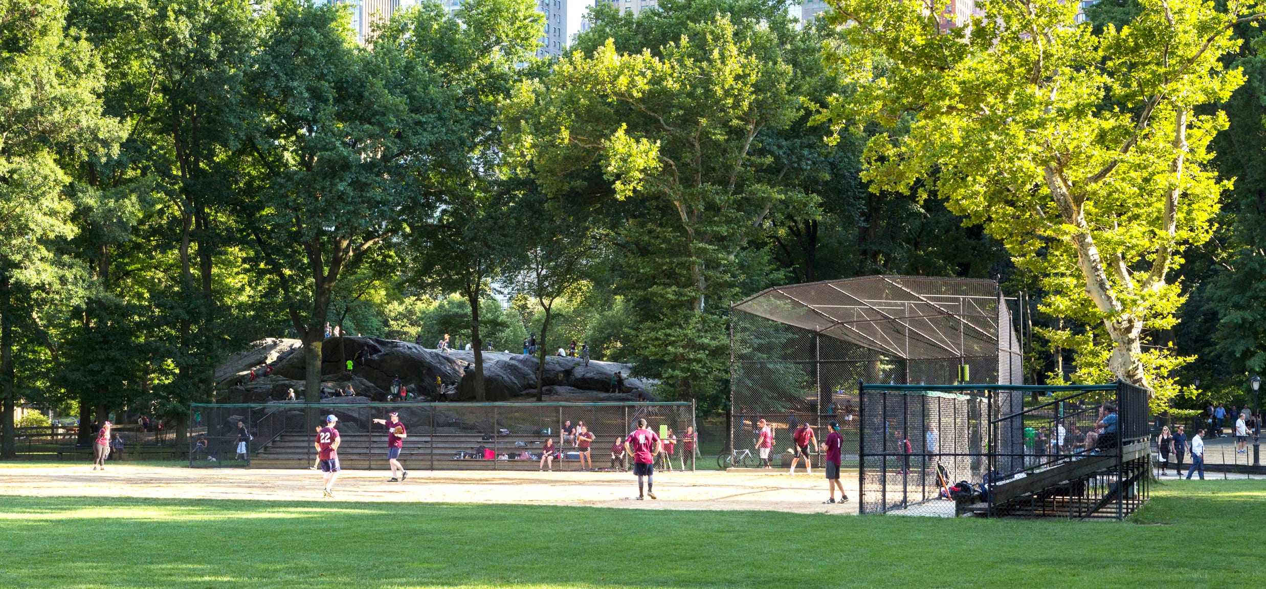 A softball game takes place under trees and the skyline of 59th Street