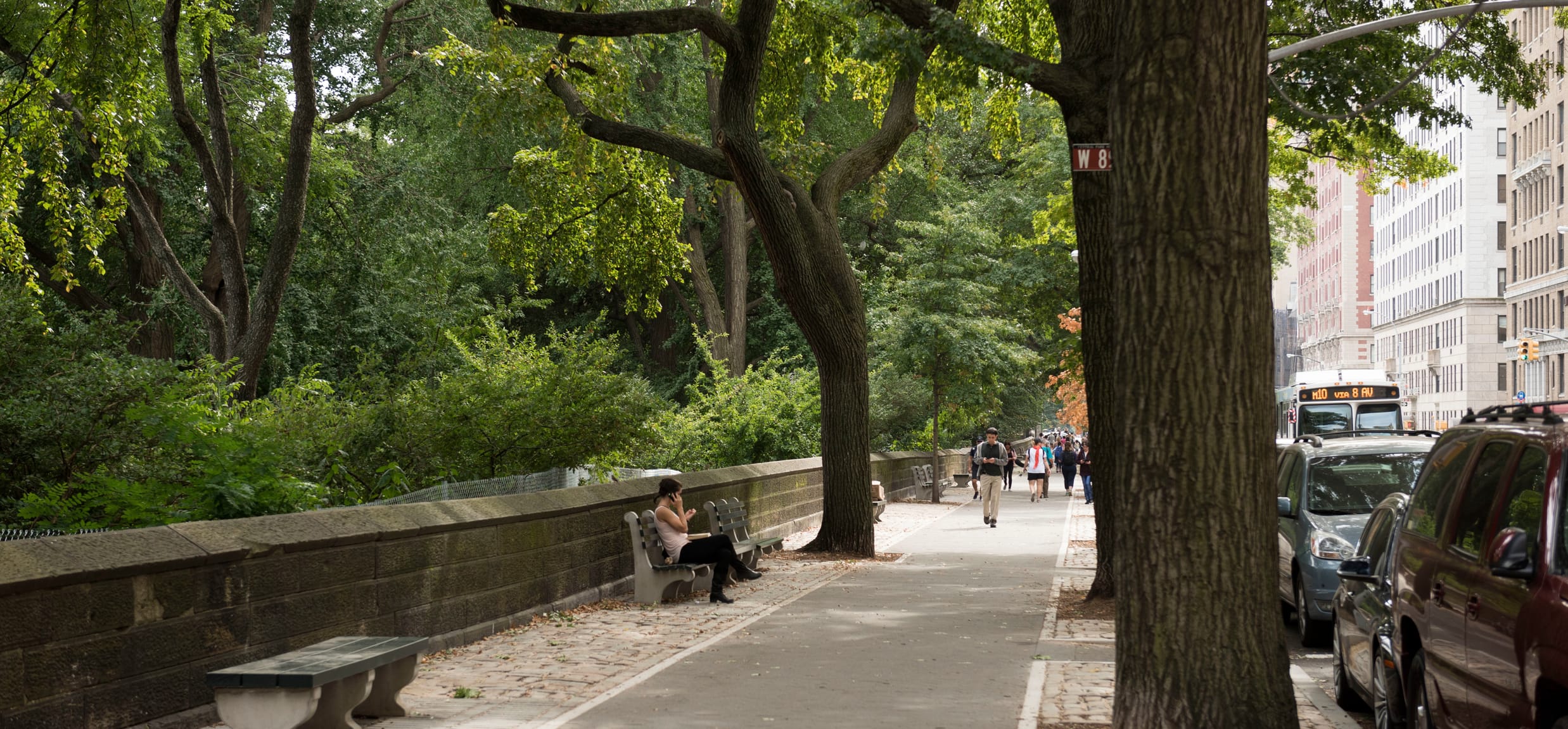 A view of the perimeter in summer, with pedestrians in the distance, one of them sitting on a park bench.