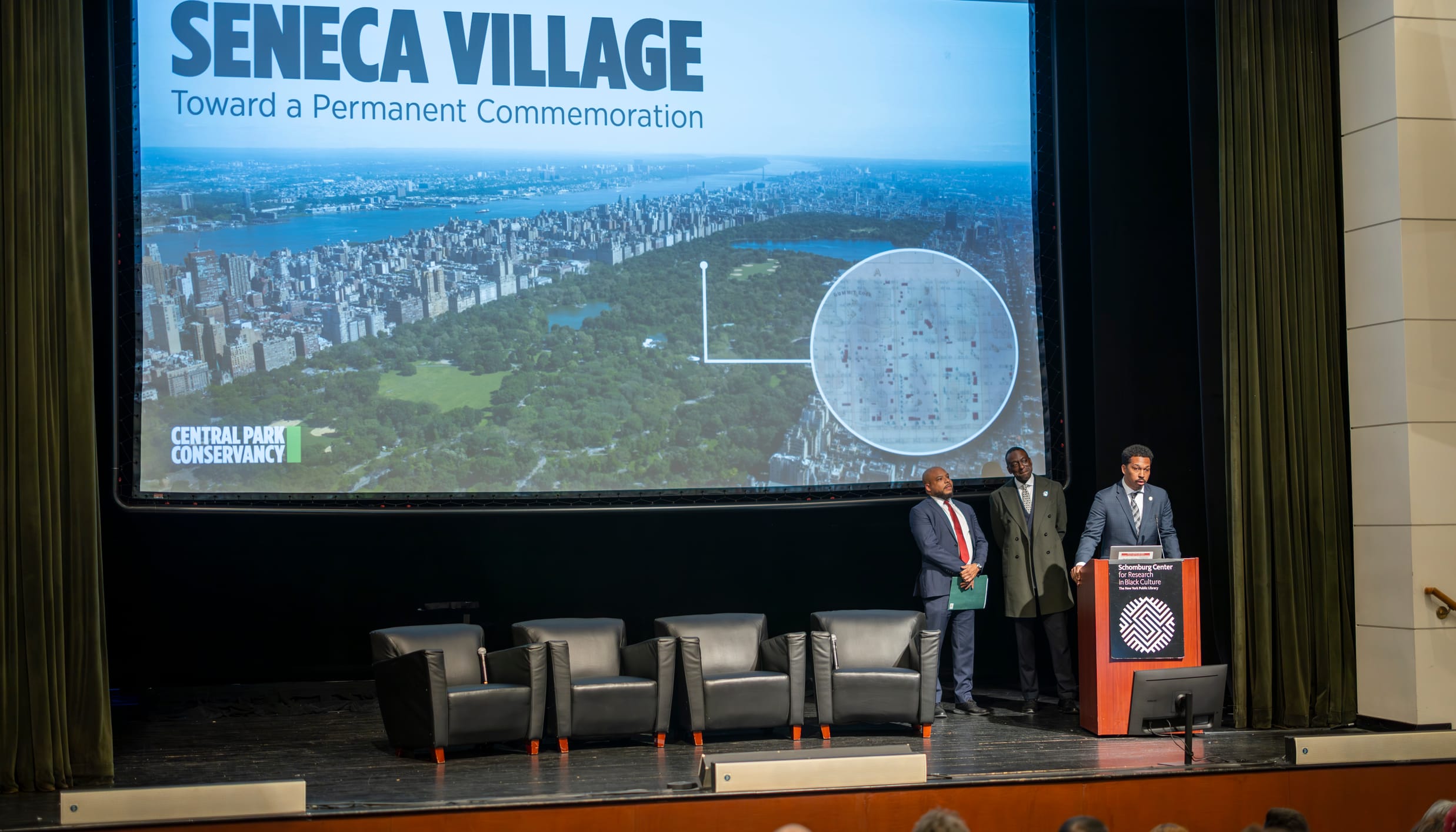 Three Black men stand at the podium at the Schomburg Center for Research in Black Culture in Harlem. On the screen behind them is a photo of Central Park with a historical map of Seneca Village.